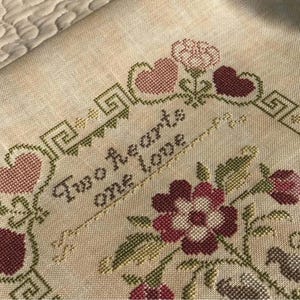 May include: Cross-stitched fabric with the words "Two hearts one love" in dark brown thread. The design includes pink hearts, flowers, and green leaves, all stitched on a beige background. The border features a geometric pattern.
