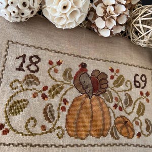 May include: Cross-stitch embroidery with a Thanksgiving theme. The design features a large orange pumpkin, a turkey, and the numbers "18" and "69". The embroidery is surrounded by green leaves and red berries, all on a beige fabric.