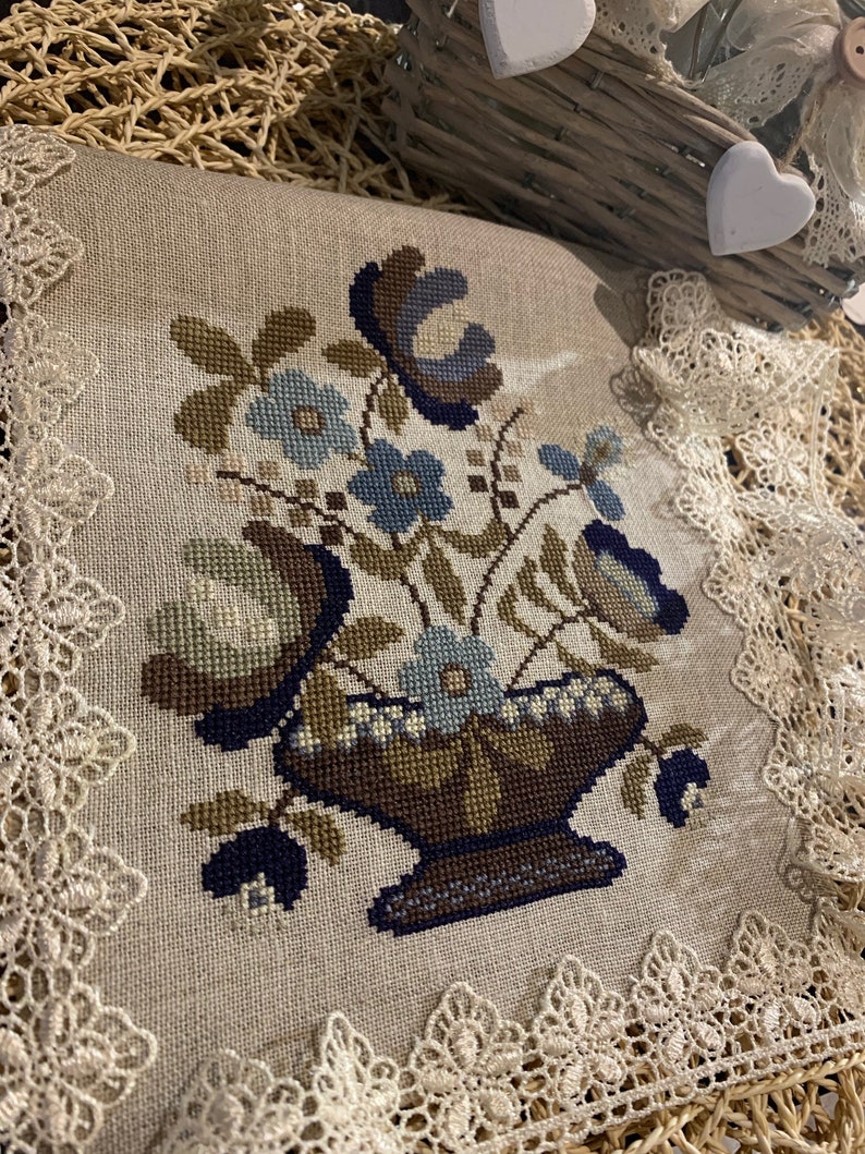 Op de afbeelding: Een beige linnen kussen met een blauw en bruin borduurwerk met bloemen. Het kussen is afgewerkt met witte kant.