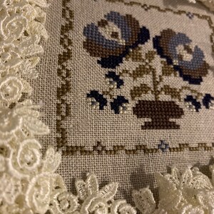 K&ouml;nnte beinhalten: Eine Kreuzstich-Stickerei mit zwei blauen Blumen in einem braunen Topf, gerahmt von wei&szlig;er Spitze.