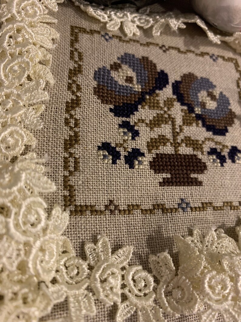 K&ouml;nnte beinhalten: Eine Kreuzstich-Stickerei mit zwei blauen Blumen in einem braunen Topf, gerahmt von wei&szlig;er Spitze.