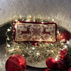 Könnte beinhalten: Ein rotes und weißes besticktes Kissen mit zwei Rentieren und einem Schneeflockenmuster. Das Kissen hat das Jahr 2024 bestickt. Das Kissen ist von einer Lichterkette und roten Kugeln umgeben.