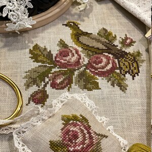 K&ouml;nnte beinhalten: Ein Kreuzstich-Stickprojekt mit einem Vogel, der auf einem Zweig mit rosa und wei&szlig;en Rosen sitzt. Das Projekt ist gerahmt und auf einem Tisch mit einer Garnrolle und einem kleinen Kissen mit &auml;hnlichem Design ausgestellt.