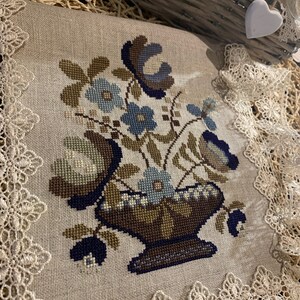 Op de afbeelding: Een beige linnen kussen met een blauw en bruin borduurwerk met bloemen. Het kussen is afgewerkt met witte kant.