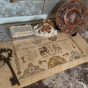 May include: A beige linen needlepoint with brown embroidery depicting keys, lions, and the text "The keys to happiness are dreams that come true".
