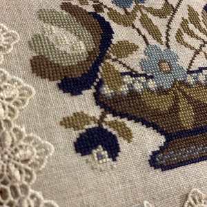 Op de afbeelding: Een close-up van een kruissteekborduursel met een bloemstuk in een bruine en blauwe vaas. Het borduursel is omlijst met een witte kant.
