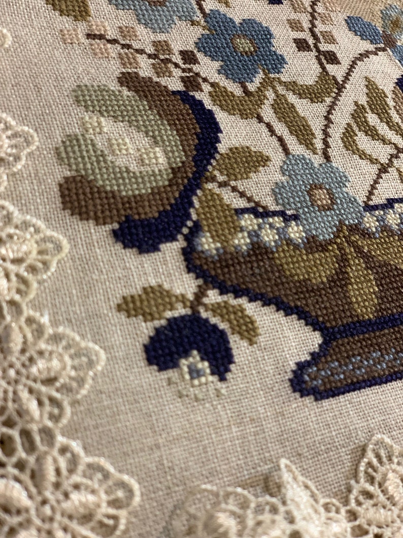 Op de afbeelding: Een close-up van een kruissteekborduursel met een bloemstuk in een bruine en blauwe vaas. Het borduursel is omlijst met een witte kant.