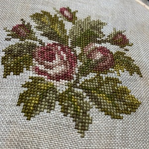 K&ouml;nnte beinhalten: Nahaufnahme einer Kreuzstich-Stickerei mit einer Gruppe von rosa und wei&szlig;en Rosen mit gr&uuml;nen Bl&auml;ttern.