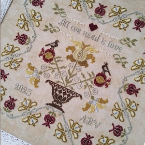 Peut inclure: Point de croix avec un motif floral. Le tissu beige présente un vase central avec des fleurs, des oiseaux, des papillons et le texte "All we need is love". Le motif incorpore du fil bordeaux, jaune et vert, avec une bordure décorative.
