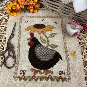 Puede incluir: Un muestrario de punto de cruz con una gallina negra y gris con un girasol, enmarcado por un borde decorativo. El muestrario incluye los números "20" y "24" y está rodeado de elementos decorativos, como unas tijeras, flores y una figura de pájaro.