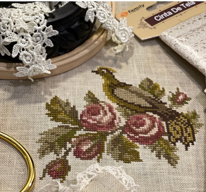 K&ouml;nnte beinhalten: Kreuzstich-Stickerei eines Vogels, der auf einem Strau&szlig; rosa Rosen mit gr&uuml;nen Bl&auml;ttern sitzt. Die Stickerei ist auf einem beigefarbenen Leinenstoff.