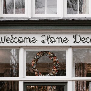 May include: A storefront with a sign that reads "Welcome Home Decor". The storefront has large windows with a fall-themed wreath in the center window.