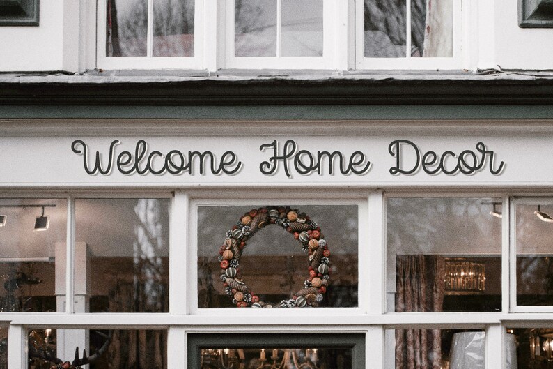 May include: A storefront with a sign that reads "Welcome Home Decor". The storefront has large windows with a fall-themed wreath in the center window.