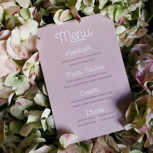 May include: A pink menu card with white text listing appetizers, main courses, desserts, and drinks. The card is lying on a bed of white and pink hydrangea flowers.