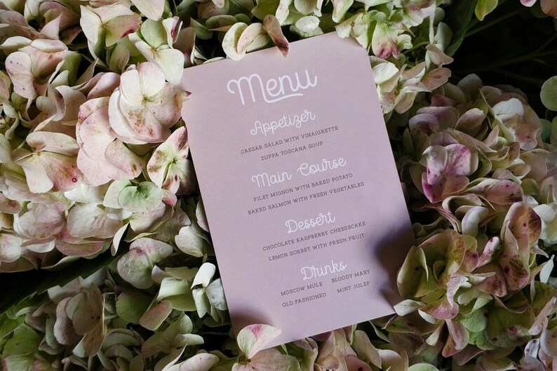 May include: A pink menu card with white text listing appetizers, main courses, desserts, and drinks. The card is lying on a bed of white and pink hydrangea flowers.
