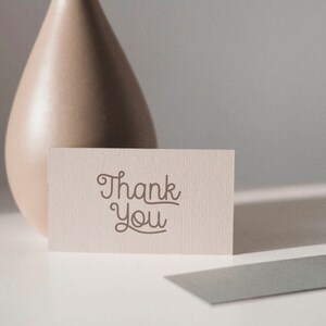 May include: A white card with the text "Thank You" written in a cursive font. The card is sitting on a white surface next to a beige ceramic vase.