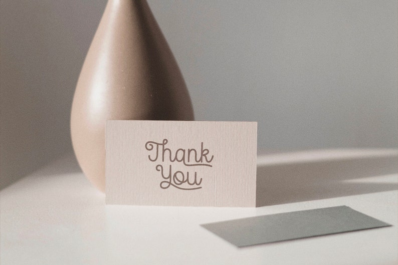 May include: A white card with the text "Thank You" written in a cursive font. The card is sitting on a white surface next to a beige ceramic vase.