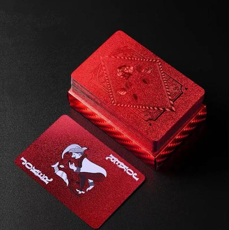 May include: A deck of red playing cards with a textured, glittery finish. One card is face up, displaying a joker illustration and the word "JOKER" in white. The deck is stacked, with a rose design on the top card. The cards are on a dark surface.