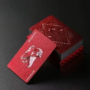 May include: A deck of red playing cards with a glittery finish. The Joker card is in the foreground, displaying a detailed illustration. The card's corners are labelled "JOKER". The deck is stacked on a dark surface.