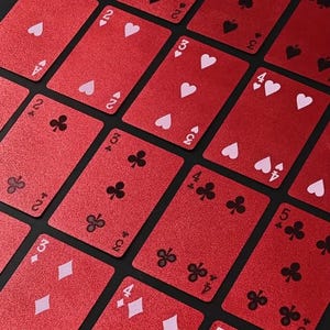 May include: A selection of red playing cards with white suits and numbers. The cards display hearts, clubs, and diamonds. The cards are arranged on a dark surface, creating a pattern. The cards are a standard size.