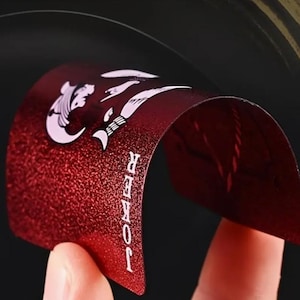 May include: A red playing card with a textured surface is bent to reveal the word "JOKER" in white text. The card features white illustrations and a curved design, set against a dark background.