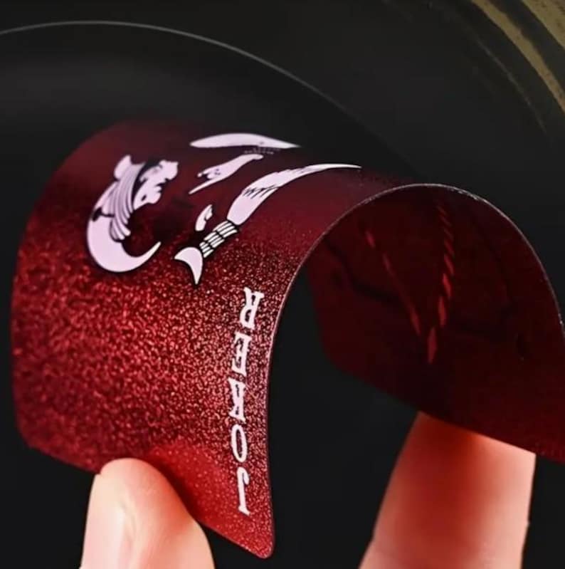 May include: A red playing card with a textured surface is bent to reveal the word "JOKER" in white text. The card features white illustrations and a curved design, set against a dark background.