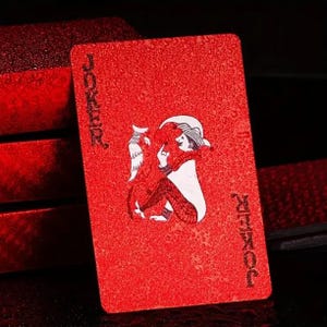 May include: A red Joker playing card with a textured surface. The card features a white-clad jester with a hat and a feather, and the word "JOKER" is printed in white on the top and bottom.