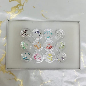 May include: A set of 12 small clear containers with colourful rhinestones inside. The containers are arranged in a circle in a white box.