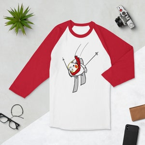May include: White and red raglan baseball tee with a cartoon illustration of a yellow cockatiel wearing a red scarf and skiing down a slope. The bird is on skis and holding ski poles. The shirt has red sleeves.