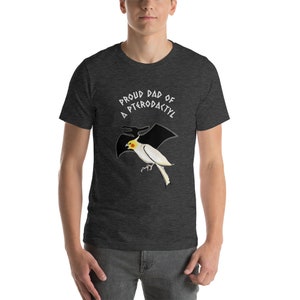 May include: Dark gray t-shirt with the text "Proud Dad of a Pterodactyl" in white. The shirt features a cartoon illustration of a white bird with a yellow head and black wings, resembling a pterodactyl.