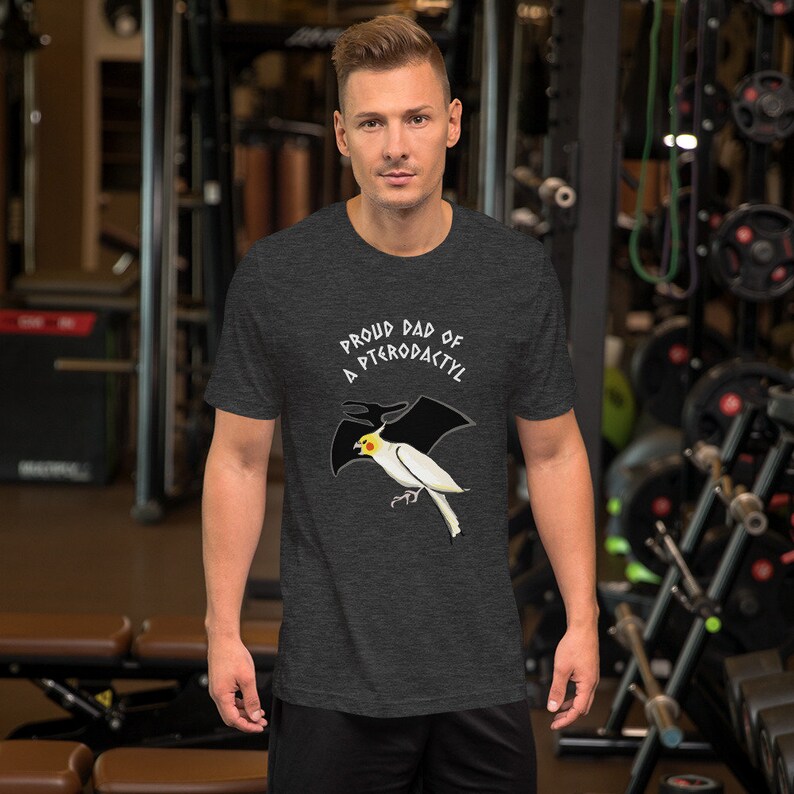 May include: Dark gray t-shirt with the text "Proud Dad of a Pterodactyl" above a cartoon illustration of a white and yellow bird with black wings. The shirt is made of a soft material.