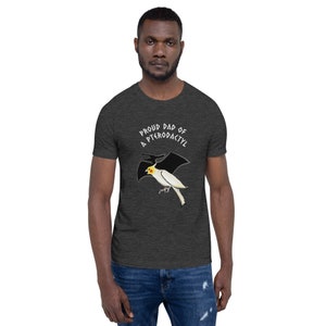 May include: Dark gray t-shirt with the text "Proud Dad of a Pterodactyl" above a cartoon illustration of a white bird with a yellow head and black wings. The bird is flying. The shirt is short-sleeved.