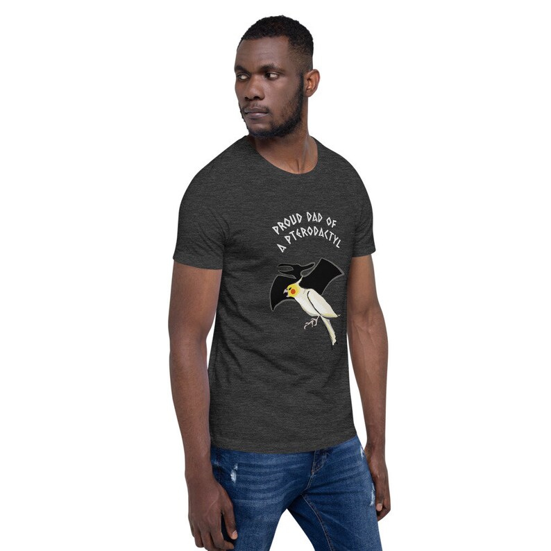 May include: Dark gray t-shirt with the text "PROUD DAD OF A PTERODACTYL" above a cartoon bird with pterodactyl wings. The bird is white with a yellow head and black wings. The shirt is made of a soft material.