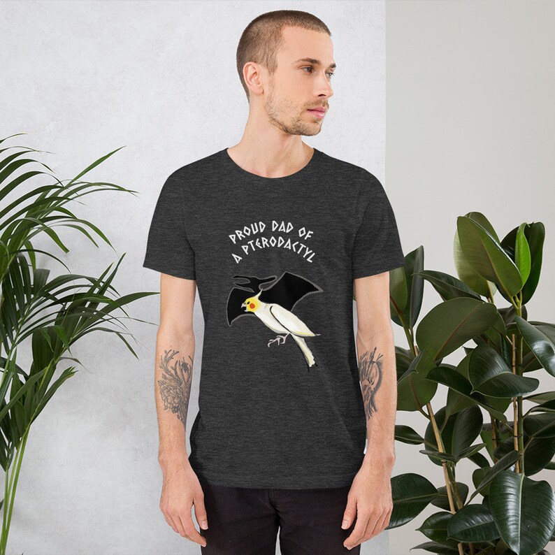 May include: Dark gray t-shirt with the text "Proud Dad of a Pterodactyl" above a cartoon illustration of a white and yellow bird with black wings. The shirt is made of a soft material and has short sleeves.