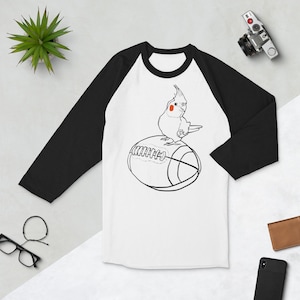 May include: White and black raglan baseball tee featuring a cartoon cockatiel perched on a football. The bird is drawn in black outline with a red cheek patch. The shirt has black sleeves and a white body.