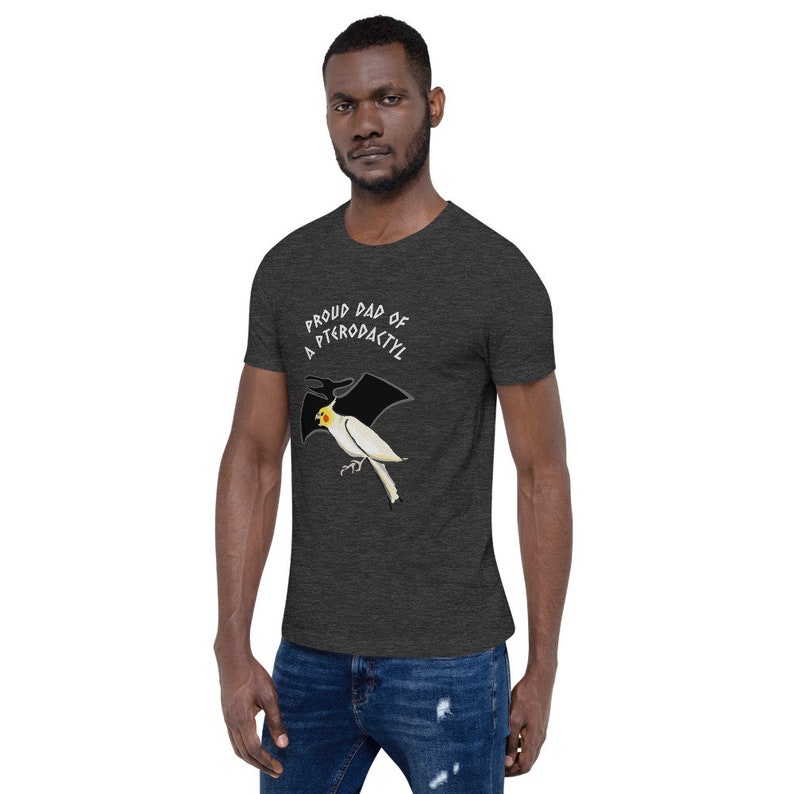 May include: Dark gray t-shirt featuring a cartoon bird with pterodactyl wings. The text "PROUD DAD OF A PTERODACTYL" is arched above the bird. The bird is white with a yellow head and black wings. The shirt is made of a soft material.