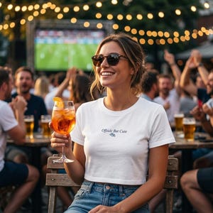 Puede incluir: Una mujer con gafas de sol y una camiseta corta blanca con el texto "Offside But Cute" y un gráfico de balón de fútbol. Sostiene un cóctel con una rodaja de naranja, sonriendo, rodeada de gente.