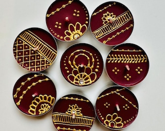May include: Seven round tea light candles with deep burgundy wax, each decorated with intricate gold paint patterns featuring floral motifs, geometric lines, and dots. Some candles include small clear rhinestones embedded in the gold designs.