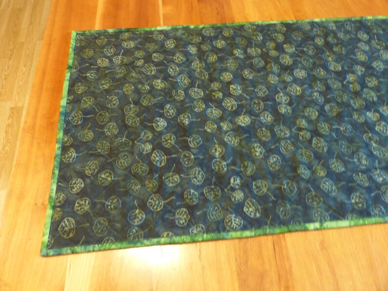 May include: A rectangular fabric table runner featuring a dark blue and black batik-style background with a repeating pattern of light green and yellow outlined leaves. The runner is finished with a bright green mottled fabric border.