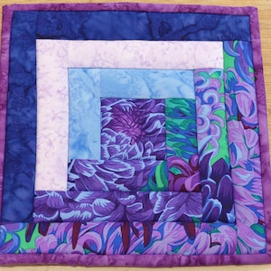 May include: Square quilted fabric coaster with a floral design in shades of purple, blue, and green. The coaster features a log cabin pattern with a purple border. The center showcases a large purple flower.
