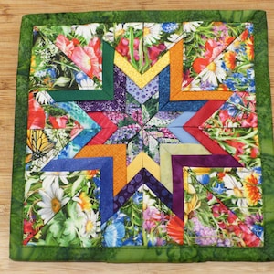 May include: A square fabric quilt block featuring a central star pattern composed of multi-colored geometric shapes. The design incorporates floral prints with daisies and poppies, alongside solid-colored fabric segments in orange, purple, blue, and yellow, all framed by a green border.