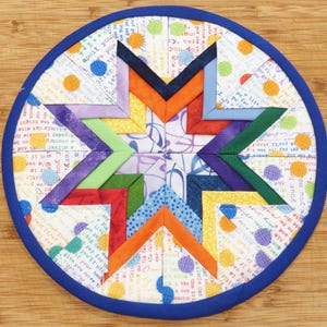 May include: A round, blue-trimmed, quilted hot pad with a colorful star design. The star is made of multiple fabric strips in shades of purple, green, blue, orange, red, and yellow. The background fabric is white with a light blue and orange polka dot pattern and text.