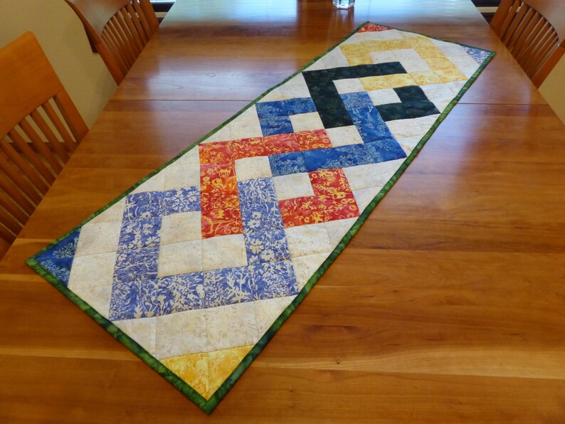 May include: A rectangular quilted table runner featuring a geometric interlocking pattern. The design consists of squares in shades of yellow, orange, blue, and dark green, set against a cream-colored background with a green fabric border.