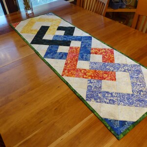 May include: A rectangular quilted table runner featuring a geometric interlocking pattern. The design incorporates fabric squares in shades of yellow, black, blue, and orange with floral and marbled textures, all bordered by a green fabric trim.