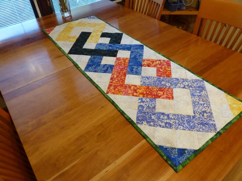 May include: A rectangular quilted table runner featuring a geometric interlocking pattern. The design incorporates fabric squares in shades of yellow, black, blue, and orange with floral and marbled textures, all bordered by a green fabric trim.