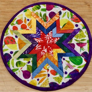 May include: A circular quilted potholder featuring a central star pattern composed of multi-colored fabric segments. The outer border is a solid purple fabric, while the inner sections display a white background with a colorful fruit and vegetable print.