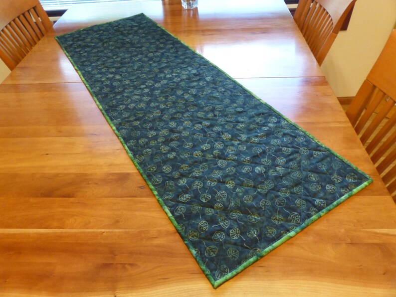 May include: A rectangular quilted table runner featuring a dark teal fabric with a repeating pattern of light green leaf outlines. The runner has a thin, bright green fabric border and diagonal stitching lines across the entire surface.