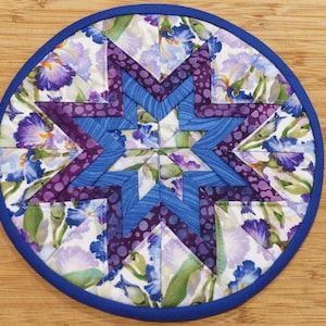 May include: A round, quilted trivet with a blue border. The center features a star design in shades of blue and purple, with floral fabric accents. The trivet is approximately 8 inches in diameter.