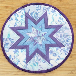 May include: A round, quilted trivet featuring a starburst design. The star is composed of various shades of blue and purple fabric, with a floral pattern on the outer ring. The trivet is approximately 8 inches in diameter.