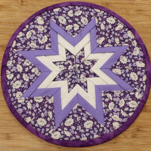 May include: Round, handmade quilted trivet featuring a star design. The trivet has a floral pattern in shades of purple and white, with a lavender star in the center. The edge is trimmed with a solid purple binding. This decorative item is suitable for kitchen use.